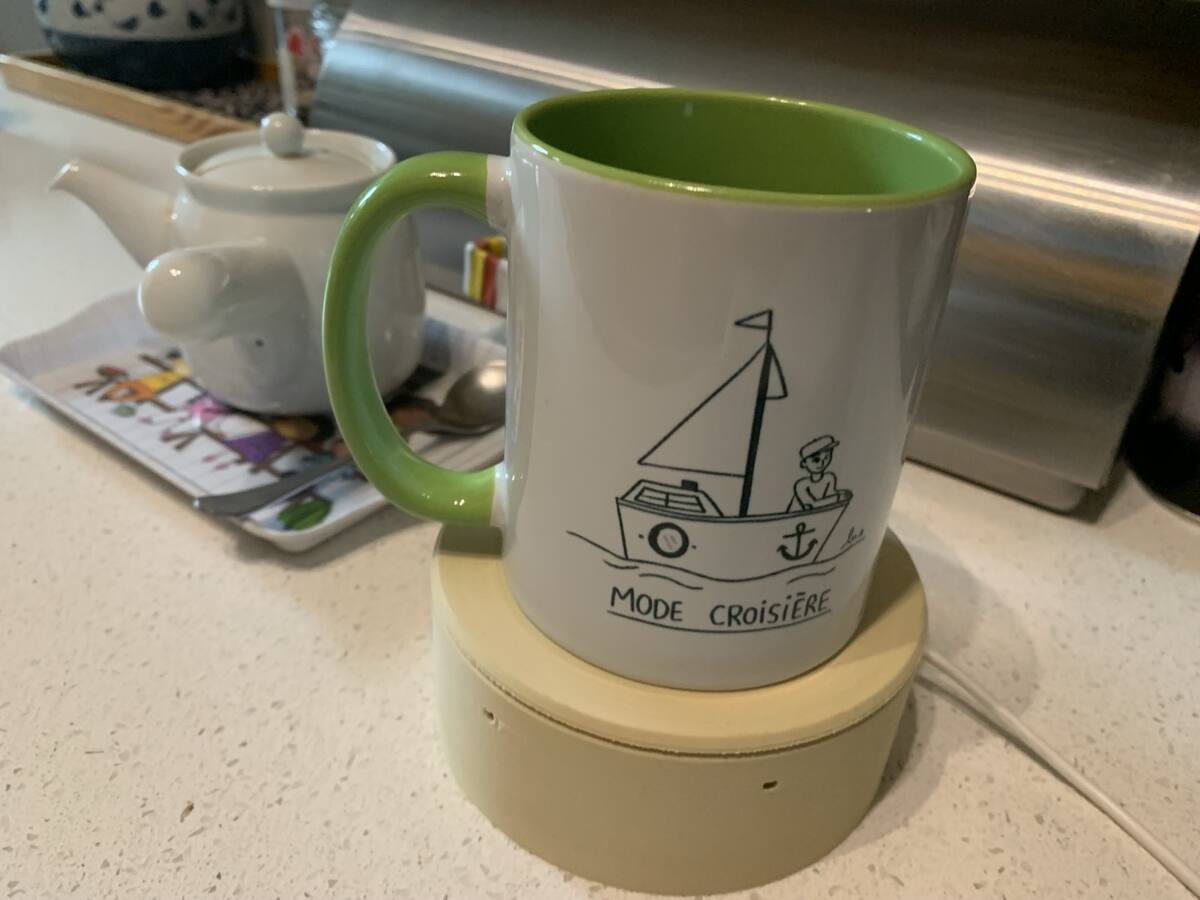 Une tasse blanche à l'intérieur vert, illustrée d'un petit bateau et titrée « Mode croisière », posée sur la Kluk, un socle rond beige, à côté d'une théière.